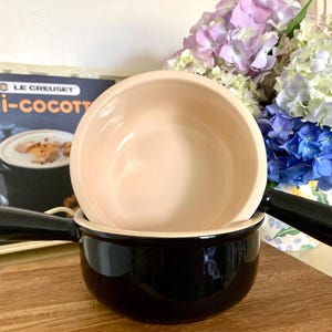 Peut inclure: Une mini-cocotte en céramique noire avec un intérieur crème et une longue poignée, dotée d'une finition lisse et brillante. Le pot est rond avec une large ouverture, conçu pour des portions individuelles, et présente un style culinaire classique.