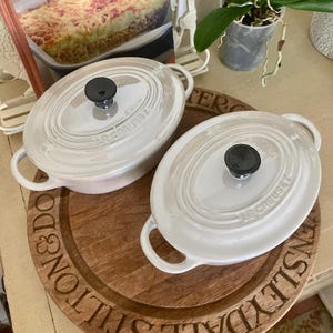 May include: Two white oval Le Creuset cocottes with black knobs and handles sit on a wooden serving board. The board has engraved text around the edge. A cookbook and a potted orchid are in the background.