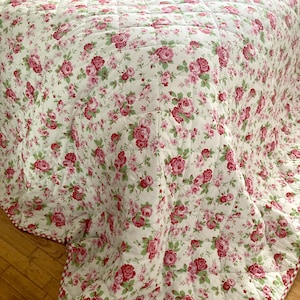May include: A quilted bedspread featuring a white background with a repeating pattern of pink roses and green leaves. The fabric is stitched in a grid pattern across the entire surface, creating a textured, soft appearance for bedding.