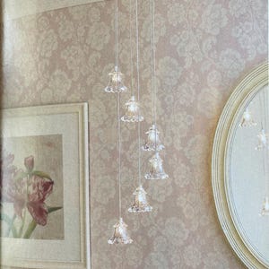 Könnte beinhalten: Ein Kronleuchter mit sechs durchsichtigen, glockenförmigen Glasschirmen hängt von der Decke. Die Schirme sind an dünnen Drähten vor einem floralen Tapetenhintergrund aufgehängt. Ein gerahmter Blumendruck und ein runder Spiegel sind ebenfalls zu sehen.