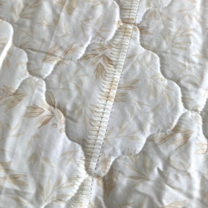 May include: Close-up of a quilted fabric with a repeating pattern of beige leaves on a white background. The fabric is stitched with a wavy pattern, and a decorative seam runs down the center.