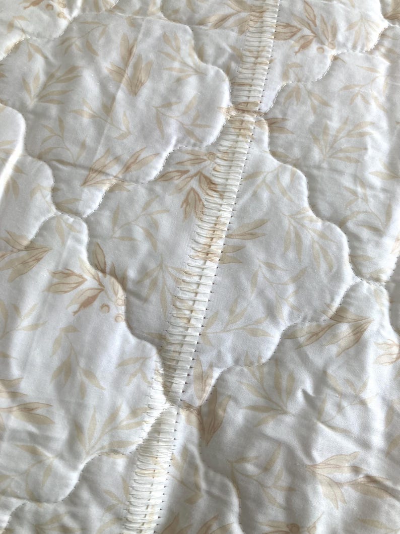 May include: Close-up of a quilted fabric with a repeating pattern of beige leaves on a white background. The fabric is stitched with a wavy pattern, and a decorative seam runs down the center.