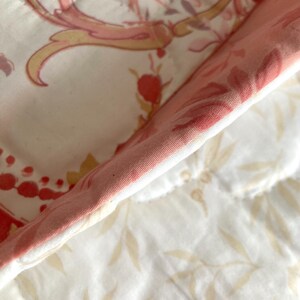 May include: Close-up of a quilted fabric with a floral pattern. The fabric features a white background with pink, red, and yellow floral and scroll designs. The edge of the fabric is a solid pink color.