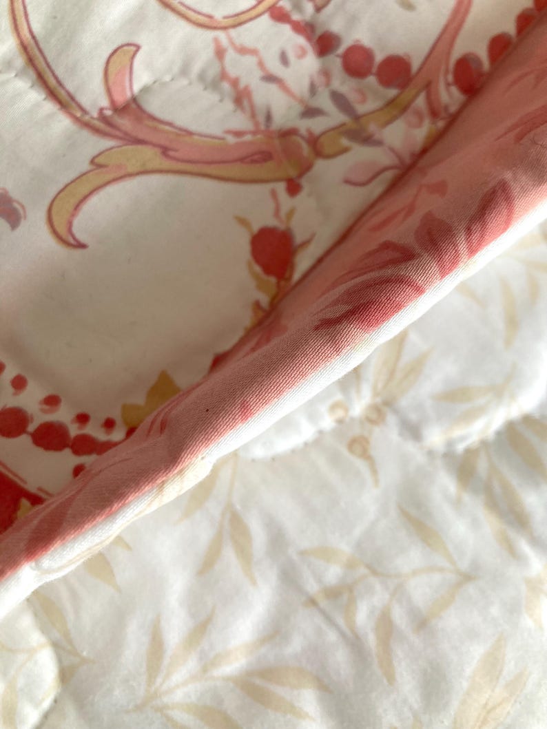 May include: Close-up of a quilted fabric with a floral pattern. The fabric features a white background with pink, red, and yellow floral and scroll designs. The edge of the fabric is a solid pink color.
