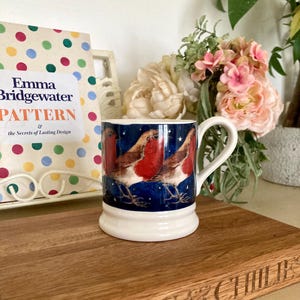 May include: A ceramic mug with a white base and handle, featuring a dark blue background with illustrations of robins. The mug is placed on a wooden surface, with a book titled "Emma Bridgewater Pattern" visible in the background.