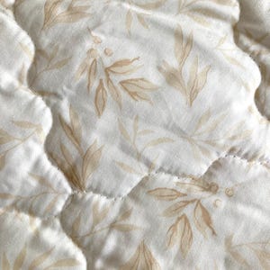 May include: Close-up of a quilted fabric with a repeating pattern of beige leaves and berries on a white background. The fabric has a wavy, quilted texture with visible stitching.