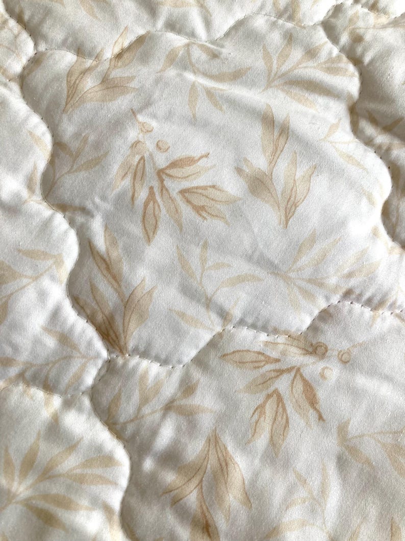 May include: Close-up of a quilted fabric with a repeating pattern of beige leaves and berries on a white background. The fabric has a wavy, quilted texture with visible stitching.