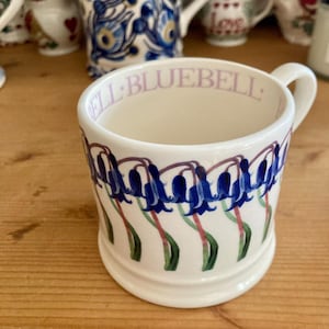 Puede incluir: Taza de cerámica blanca con un diseño floral de campanillas. La taza presenta un patrón repetido de flores azules en forma de campana con hojas verdes y tallos rojos. La palabra "BLUEBELL" está impresa en lila claro alrededor del borde.