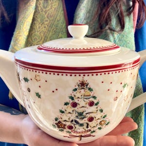 Può includere: Una teiera in ceramica color crema con un motivo natalizio a forma di albero composto da biscotti e ornamenti. La teiera presenta un coperchio abbinato con bordo rosso, un manico curvo e un beccuccio, decorata con piccole foglie di agrifoglio.