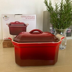 Może przedstawiać: Czerwona ceramiczna terrina z pasującą pokrywką i uchwytem. Terrina ma prostokątny kształt z zaokrąglonymi rogami i jasną obwódką. Na pudełku z tyłu terriny znajduje się napis "Terrine à foie gras et sa presse".