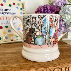 May include: A white ceramic mug featuring a colorful illustration of anthropomorphic animals cooking in a kitchen. The scene depicts a brown animal and a grey rabbit stirring a blue pot of green vegetables, with shelves of dishes in the background.