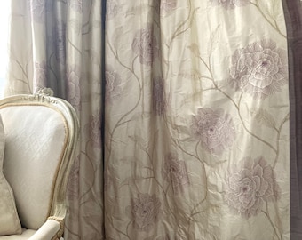 May include: Cream-colored floral curtains with large, detailed flower patterns in a muted mauve color. The curtains are draped, and a portion of a white, ornate chair is visible on the left side of the image. The fabric has a subtle sheen.
