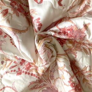 May include: White quilted fabric with a repeating floral pattern in shades of pink and peach. The design includes stylized flowers, leaves, and ribbons. The fabric is folded, creating shadows and texture.