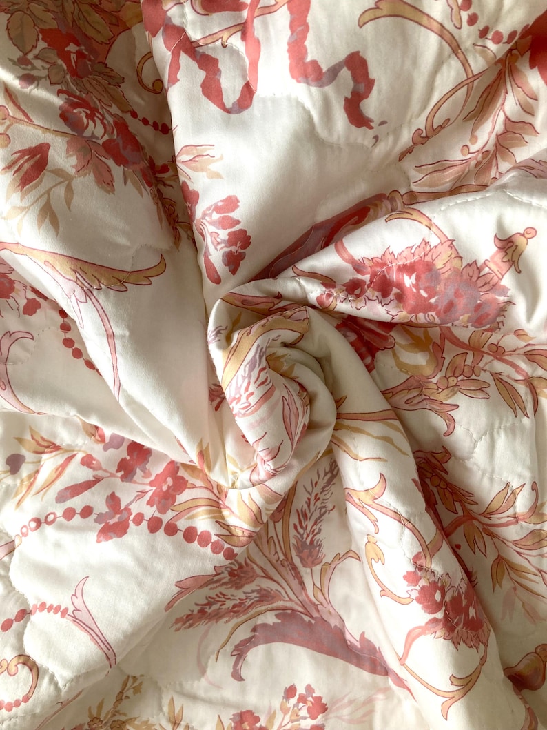 May include: White quilted fabric with a repeating floral pattern in shades of pink and peach. The design includes stylized flowers, leaves, and ribbons. The fabric is folded, creating shadows and texture.