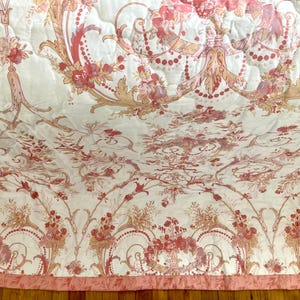 May include: A quilted bedspread with a floral pattern in shades of pink, peach, and gold on a white background. The design features ornate floral arrangements, scrolls, and decorative elements. The bedspread has a pink border.