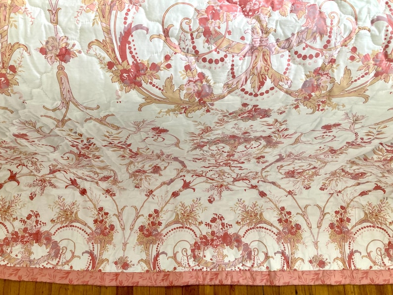 May include: A quilted bedspread with a floral pattern in shades of pink, peach, and gold on a white background. The design features ornate floral arrangements, scrolls, and decorative elements. The bedspread has a pink border.