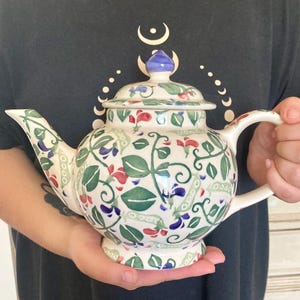Könnte beinhalten: Eine Keramik-Teekanne mit cremefarbener Basis und einem handgemalten Muster aus grünen Blättern, Ranken sowie kleinen roten und violetten Blüten. Die Teekanne besitzt einen passenden Deckel mit blauem Knauf sowie einen geschwungenen Griff.