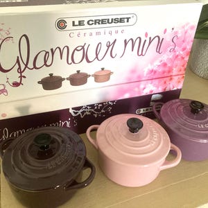 Könnte beinhalten: Ein Set aus Le Creuset Glamour Mini Keramik-Cocottes in Pflaume, Rosa und Lavendel. Jeder Mini-Topf hat einen schwarzen Knopf und zwei Griffe. Auf der Schachtel steht "Glamour mini's" und ein Blumenmuster.