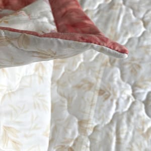 May include: Close-up of a quilted blanket with a floral pattern. The blanket features a white background with a subtle leaf design and a contrasting red border. The stitching is visible, adding texture.