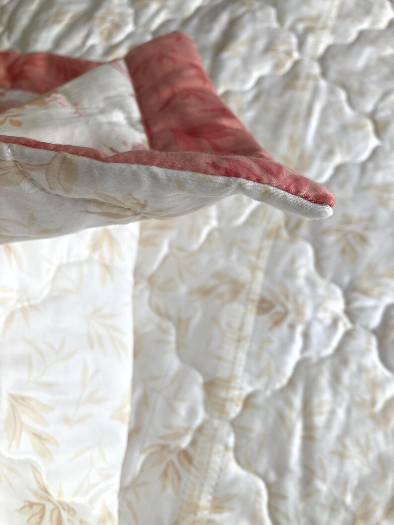 May include: Close-up of a quilted blanket with a floral pattern. The blanket features a white background with a subtle leaf design and a contrasting red border. The stitching is visible, adding texture.
