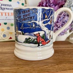 May include: A white ceramic mug featuring a blue and white winter landscape illustration with a red fox in the foreground, a tree with birds, and a church. The text A YEAR IN THE COUNTRY is printed in blue on the upper portion of the mug design.