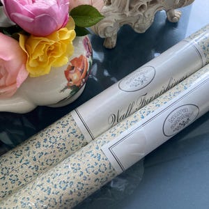 Op de afbeelding: Twee rollen Laura Ashley behang met een crèmekleurige achtergrond en een blauw bloemenpatroon. De rollen zijn gelabeld met "Wall Furnishings" en "Laura Ashley". Een vaas met roze en gele rozen staat op de achtergrond.