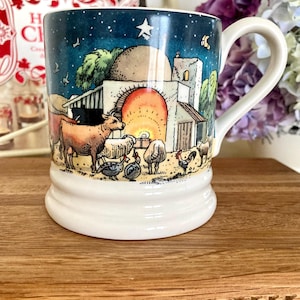 May include: A white ceramic mug featuring a colorful illustration of a nativity scene at night. The design includes a stable with a glowing orange archway, a star in the dark blue sky, and various farm animals including a cow, sheep, and rooster.