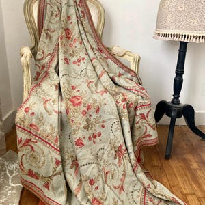 Könnte beinhalten: Eine große, rechteckige Wohndecke mit hellsalbeigrüner Grundfarbe und einem wiederkehrenden Muster aus roten Rosen, beigen Blumenranken und verzierten Schnörkelbordüren. Der Stoff hat eine weiche Textur und liegt über Rückenlehne und Sitzfläche.