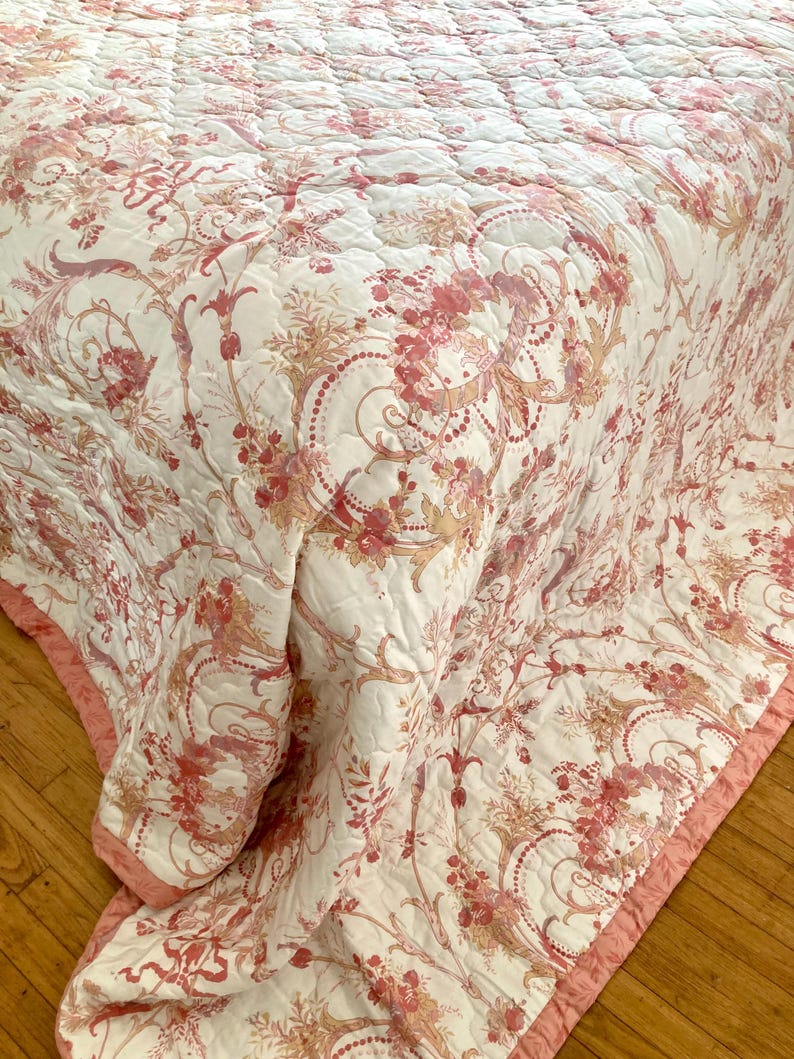 May include: A white and pink floral patterned quilt with a pink border. The quilt features a detailed design of flowers and swirling patterns. The bedding is folded over a wooden surface.