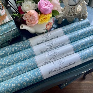 Può includere: Diversi rotoli di carta da parati floreale blu. Ogni rotolo è etichettato "Wall Furnishings". La carta da parati presenta uno sfondo azzurro con un delicato motivo floreale bianco. I rotoli sono disposti su una superficie scura, con fiori sullo sfondo.