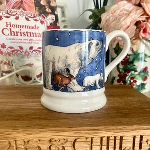 May include: White ceramic mug with a blue and white winter scene featuring a polar bear, a baby polar bear, and a brown hare. The mug has a white handle and a white base. The mug is on a wooden surface.