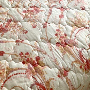 May include: Close-up of a quilted bedspread with a floral pattern. The design features red and pink flowers, leaves, and stems on a cream-colored background. The quilting creates a textured, raised effect, enhancing the intricate floral details.