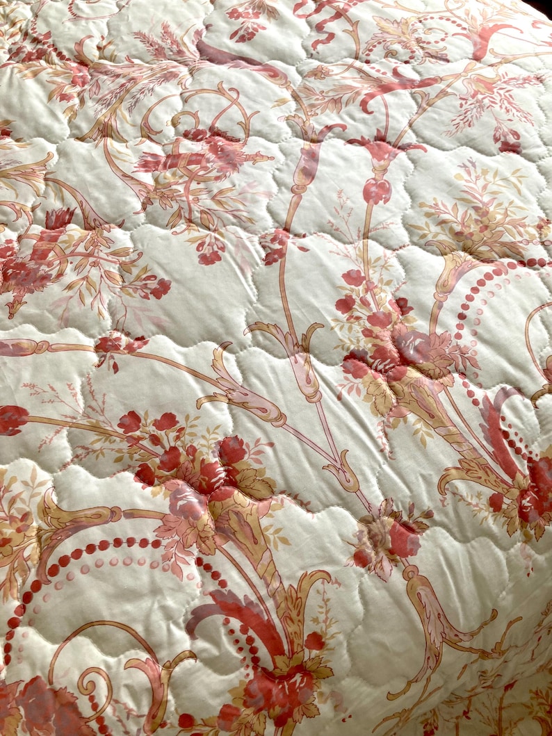 May include: Close-up of a quilted bedspread with a floral pattern. The design features red and pink flowers, leaves, and stems on a cream-colored background. The quilting creates a textured, raised effect, enhancing the intricate floral details.