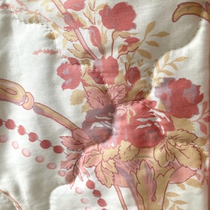 May include: Close-up of a quilted fabric with a floral pattern. The design features bouquets of red and pink roses, with yellow and peach-colored accents, set against a cream-colored background. The quilting creates a subtle, textured effect.