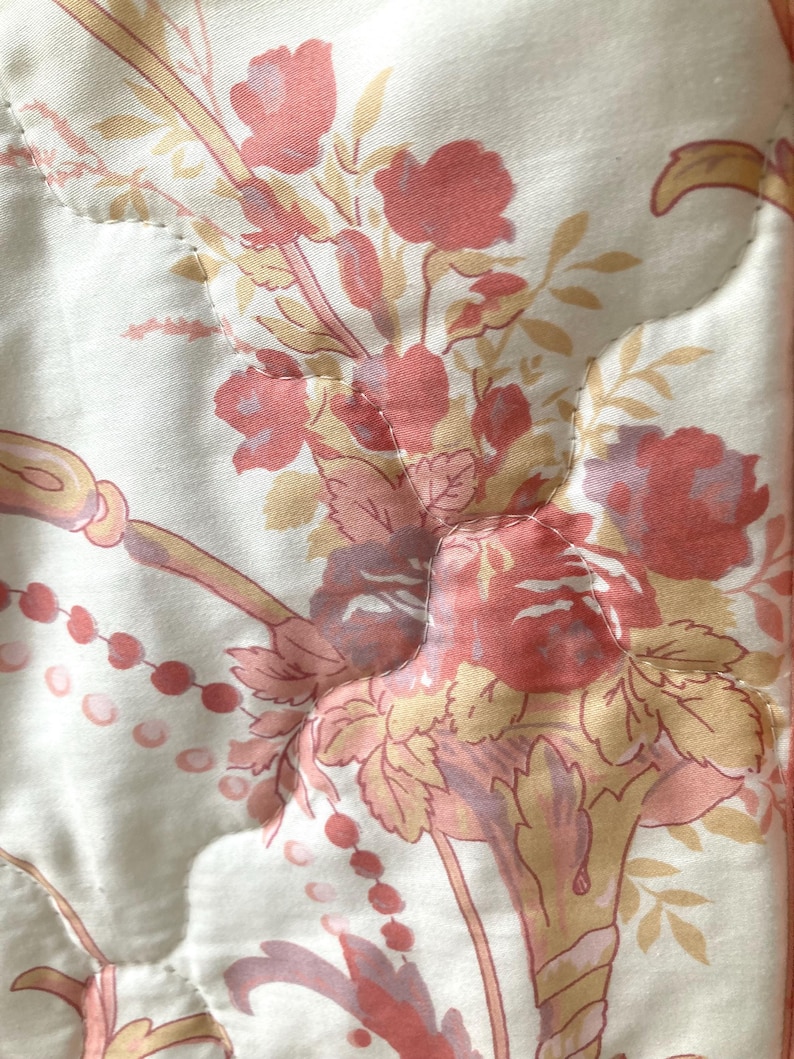 May include: Close-up of a quilted fabric with a floral pattern. The design features bouquets of red and pink roses, with yellow and peach-colored accents, set against a cream-colored background. The quilting creates a subtle, textured effect.