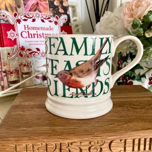 Könnte beinhalten: Ein cremefarbener Keramikbecher mit einer detaillierten Illustration eines Rotkehlchens und dem grünen Schriftzug "FAMILY FEASTS FRIENDS" auf der Vorderseite. Der Becher hat einen stabilen Henkel und einen breiten Boden in klassischem Design.