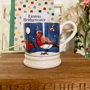 May include: A ceramic mug with a white handle and base, featuring a blue background with a robin bird design. The mug has a white moon and stars. The mug is sitting on a wooden surface.