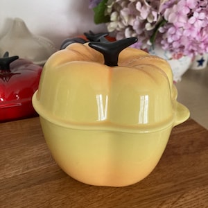 Könnte beinhalten: Eine gelbe Keramikschale in Kürbisform mit schwarzem Stiel und Deckel. Die Schale steht auf einer Holzoberfläche. Im Hintergrund ist eine rote Kürbisschale zu sehen.