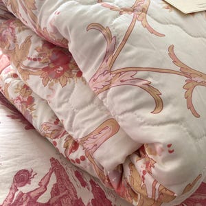 May include: A folded white and pink floral quilt with a quilted pattern. The quilt features a repeating design of pink flowers and gold-colored vines. The quilt is folded over a bedspread with a red and white toile pattern.
