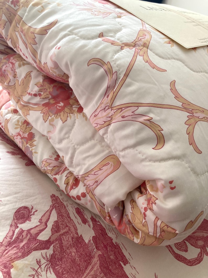 May include: A folded white and pink floral quilt with a quilted pattern. The quilt features a repeating design of pink flowers and gold-colored vines. The quilt is folded over a bedspread with a red and white toile pattern.