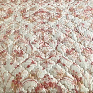 May include: A quilted bedspread with a floral pattern in shades of red, peach, and cream. The design features stylized flowers and foliage, creating a classic, elegant look. The fabric has a subtle sheen, adding to its luxurious feel.