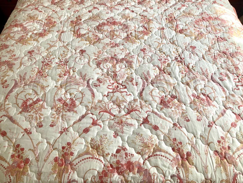 May include: A quilted bedspread with a floral pattern in shades of red, peach, and cream. The design features stylized flowers and foliage, creating a classic, elegant look. The fabric has a subtle sheen, adding to its luxurious feel.