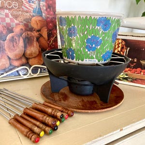 Può includere: Un set per fonduta vintage con una pentola Le Creuset a motivi floreali, un supporto in metallo nero e forchette per fonduta con manico in legno. Il set include una base in legno e un libro di cucina. La pentola ha un manico bianco.