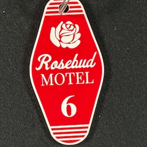 Könnte beinhalten: Ein roter Motel-Schlüsselanhänger mit weißem Rand und einem Metallring. Der Schlüsselanhänger zeigt die Worte "Rosebud MOTEL" und die Nummer "6" in Weiß. Oben befindet sich eine weiße Rosen-Grafik.
