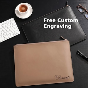 May include: Two leather zip-up document cases, one black and one tan, with the text "Free Custom Engraving" on the black case. The tan case has the name "Clemente" printed on it. A cup of coffee and a keyboard are in the background.