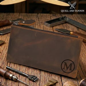 May include: A brown leather zippered pouch with the letter "M" in a circle. The pouch has a leather pull tab and the text "QUILL AND TANNIN" is visible. Various leather working tools are scattered around the pouch on a wooden surface.