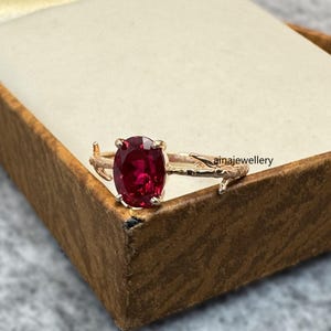 May include: A gold ring with a large, oval-shaped, deep red gemstone. The ring's band has a twig-like design. The ring is displayed inside a brown jewelry box with a cream-colored interior. The text "ainajewellery" is visible on the ring.