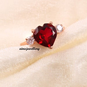 Peut inclure: Bague en or rose ornée d'une pierre rouge foncé en forme de cœur, flanquée de deux petites pierres rondes et transparentes. La bague est posée sur un fond blanc cassé. Le texte "ainajewellery" est visible.