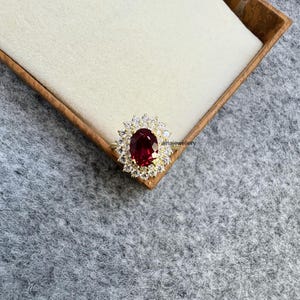 May include: A gold ring featuring a large, oval-cut, deep red gemstone surrounded by smaller, clear gemstones. The ring is set in a gold band and is displayed in a wooden box with a light-colored interior. The ring is a piece of jewelry.