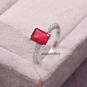 K&ouml;nnte beinhalten: Ein silberner Ring mit einem gro&szlig;en, rechteckigen, leuchtend roten Edelstein in der Mitte, flankiert von kleineren, klaren, funkelnden Steinen, die entlang der Ringschiene eingelassen sind. Der Text ainajewellery ist nahe der Basis sichtbar.